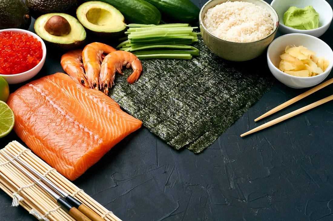 Ingredientes para sushi dispostos sobre bancada escura: filé de salmão, camarões, folhas de alga nori, arroz, pepino, abacate, gengibre e wasabi